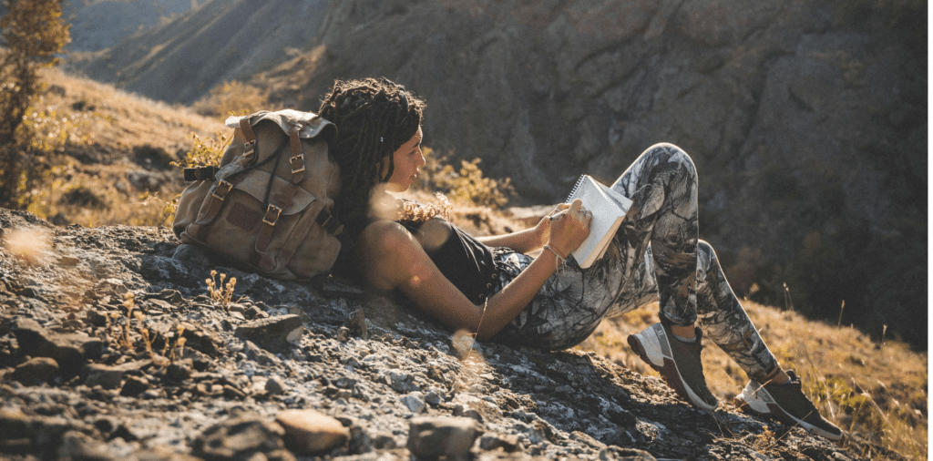 Woman laying down and journaling during a hike in the mountains. Her head is resting on her backpack and her journal rests against her legs. The scenery is rocky, with yellow grass and autumn trees.