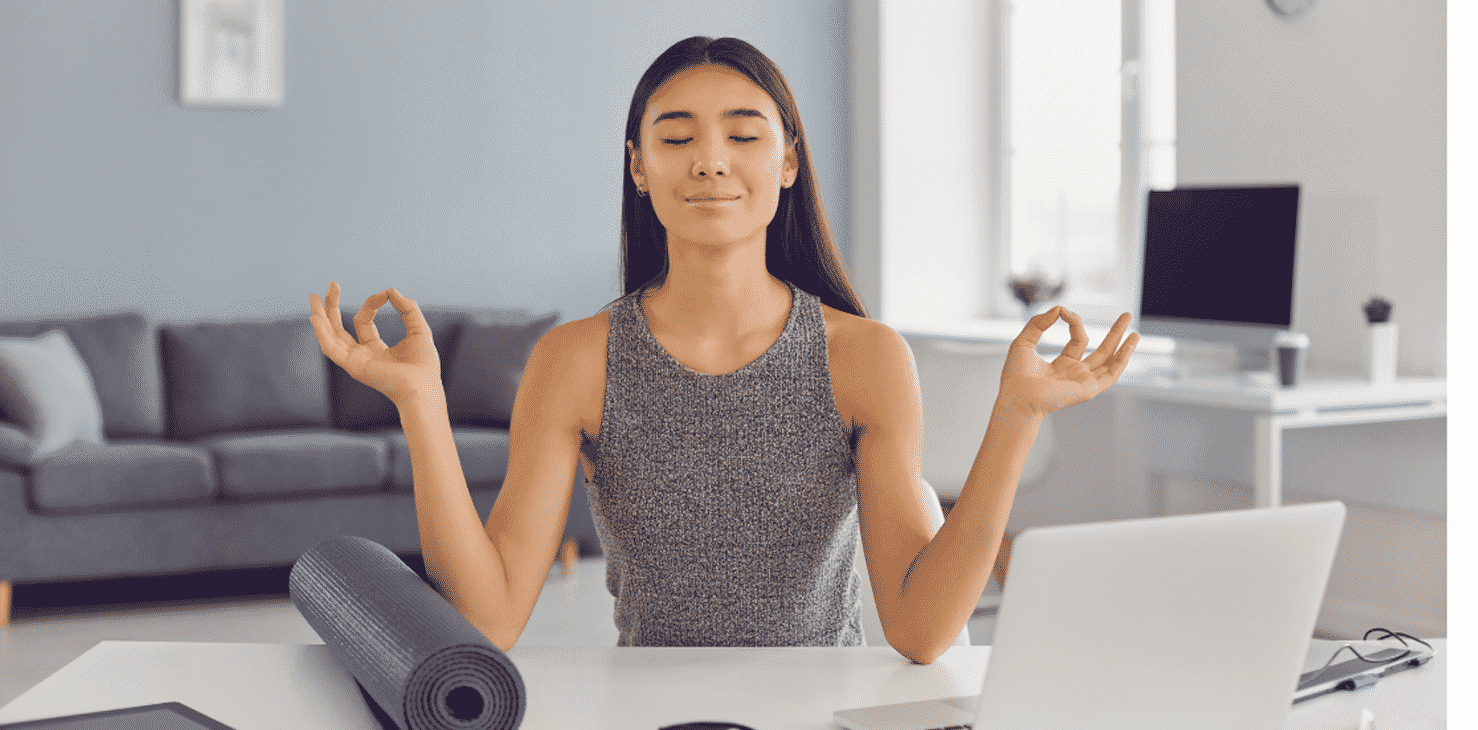 Woman meditating at her desk beside a laptop and a rolled yoga mat