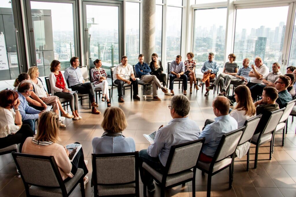 Large circle of people seated in chairs in a bright room overlooking a city