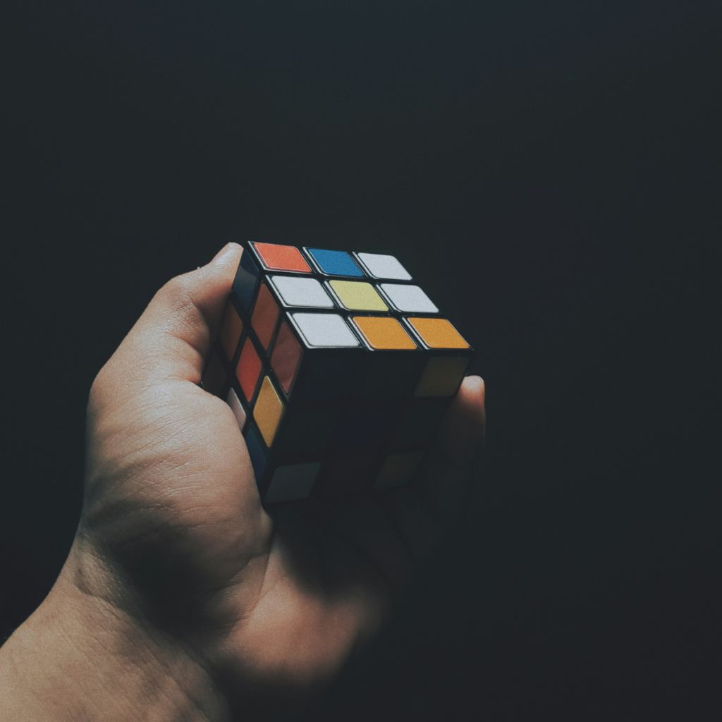 A hand holds a partially solved Rubiks Cube against a dark background, showing mixed red, blue, yellow, orange, and white squares.