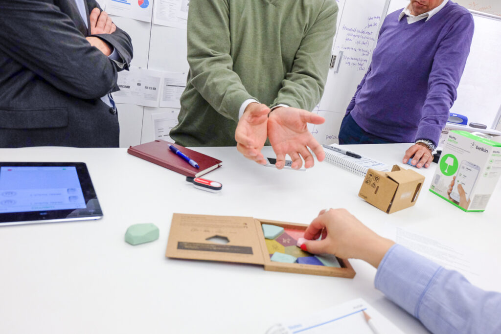 Three people stand around a white table discussing and gesturing, with hands visible. On the table are an iPad, documents, a cardboard box, a pen, and a geometric puzzle being interacted with by a fourth person.