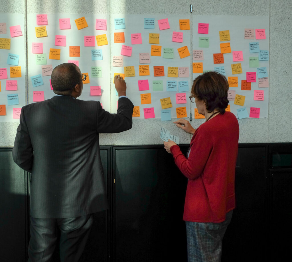 Two people stand in front of a wall covered with colorful sticky notes, reading and writing on the notes. The scene suggests brainstorming or collaborative planning in a professional setting.